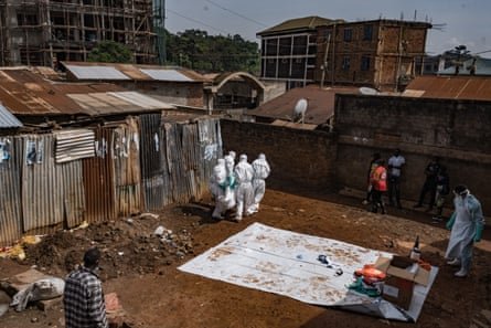 Four people in white hazmat suits carry a body out of a clearing in a shantytown as others look on
