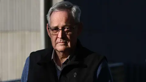 EPA Ian Wilkinson with white hair and glasses looks at the camera. He wears a blue shirt and a black vest.