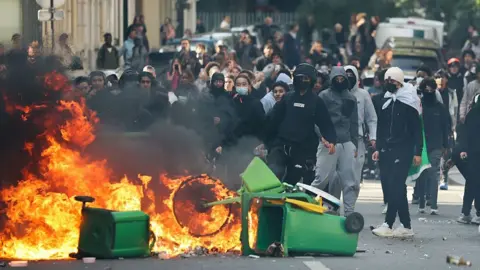 AFP via Getty Images A student sets fire to rubbish bins as protesting students carry out blockading action near place de la Nation, in Paris, on October 2, 2026. Since late September, students have staged protests outside schools over grievances ranging from timetables, class sizes and a shortage of teachers to overheated classrooms during this year's heatwaves. The protests started in the Ile-de-France region around Paris but have now spread to other cities nationwide.