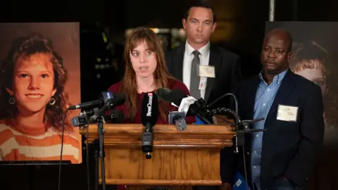 The Tennessean/Reuters A woman with lonb brown hair flowing over a burgundy blouse stands in front of a podium outfitted with multiple microphones, two suited men standing behind her, as they are flanked with enlarged photos of a blonde and smiling teenager, Colleen Slemmer, who was murdered at age 19 by Christa Pike